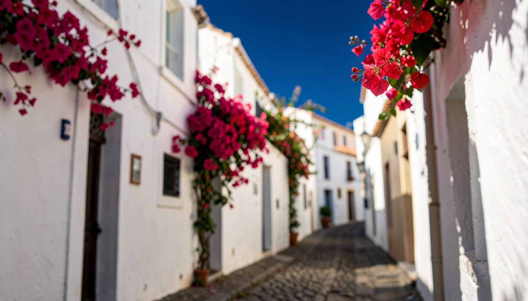 Warm Mediterranean sunlight falling across a narrow cobbled street lined with whitewashed buildings and trailing bougainvillea