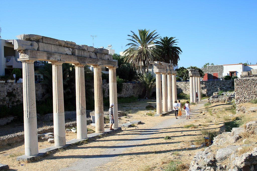 Ancient stone column ruins lining a pathway with palm trees and a stone wall under a clear blue sky