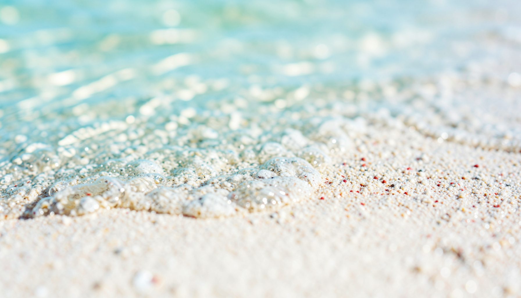 Extreme close-up of calm shallow water lapping over fine pale sand, soft ripples catching sunlight in shades of aquamarine