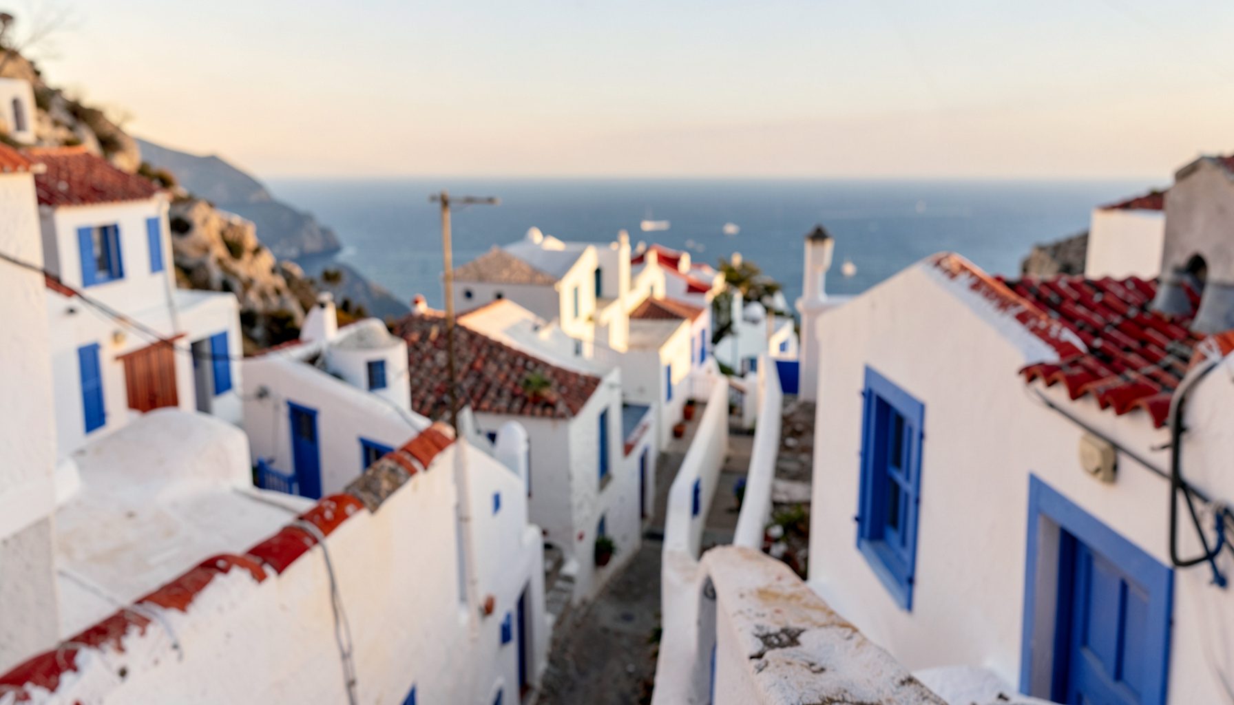 Whitewashed hillside village houses with blue shutters, narrow lanes, and distant sea view in warm afternoon light