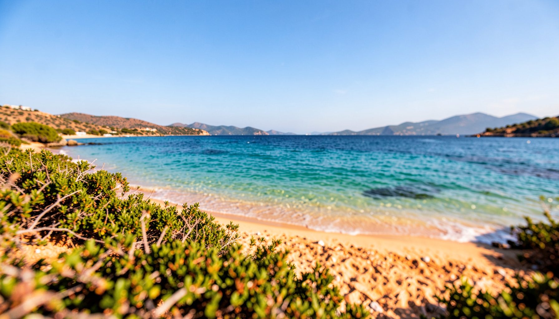 Wide panorama of the Aegean Sea under a bright blue sky, with calm turquoise water meeting a golden sandy shoreline, framed by distant hills